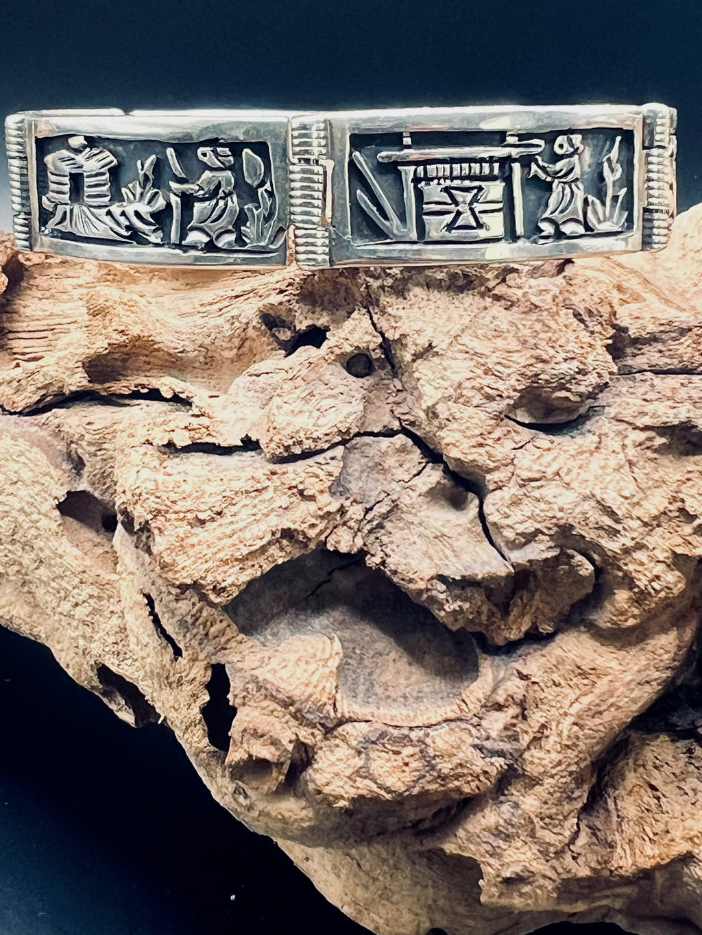 Sterling silver story panel bracelet depicting a woman weaving a blanket on a traditional loom and a woman walking through the desert towards a hut. The bracelet sits on a gnarled vine.